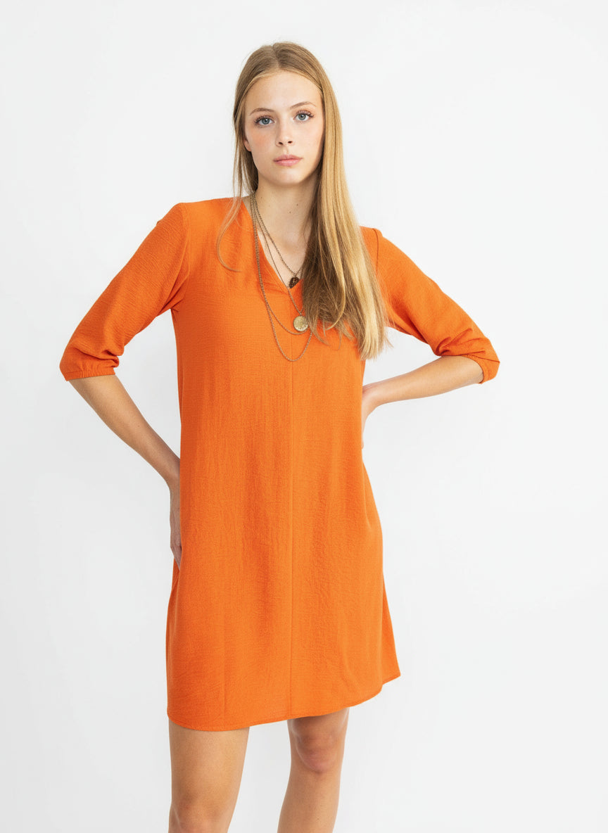 A woman standing in a studio setting, wearing a orange mini dress with a V-neck and elastic sleeves.
