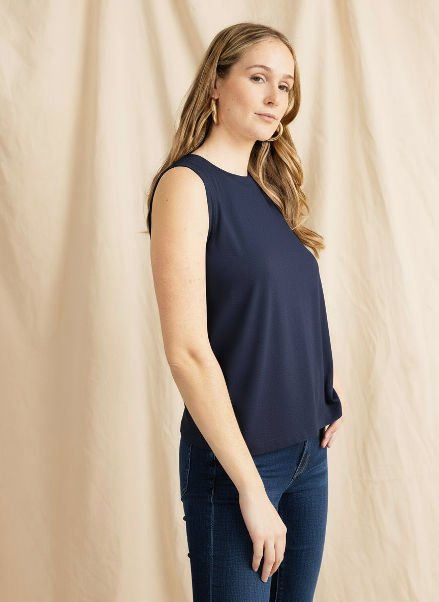Woman wearing a navy sleeveless top and blue jeans against a beige curtain.
