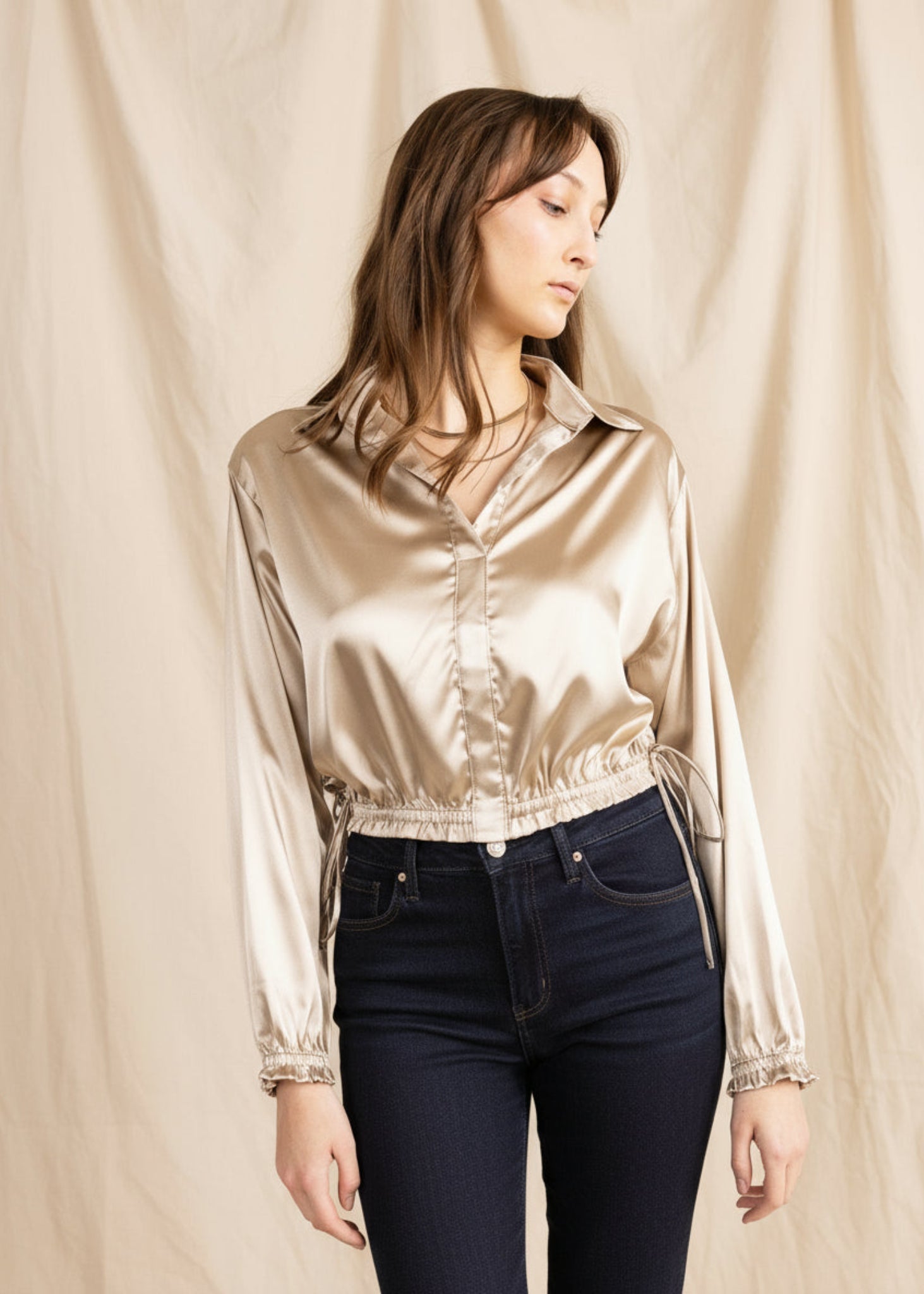 Woman wearing a shiny gold blouse and blue jeans against a beige background