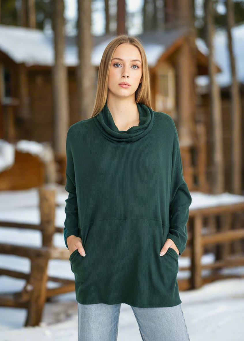 Woman wearing a green cowl-neck sweater and cap in a snowy outdoor setting with wooden structures.
