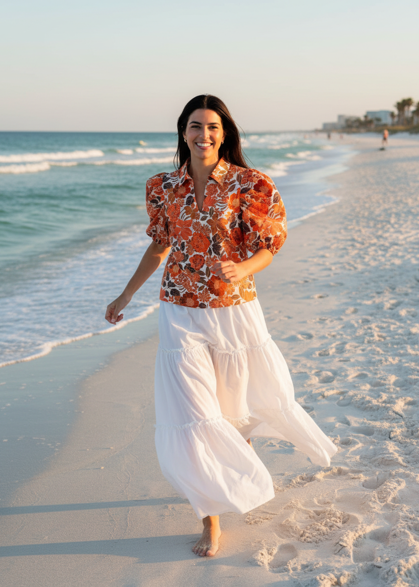 Woman in a floral shirt and white skirt walking on a beach.
