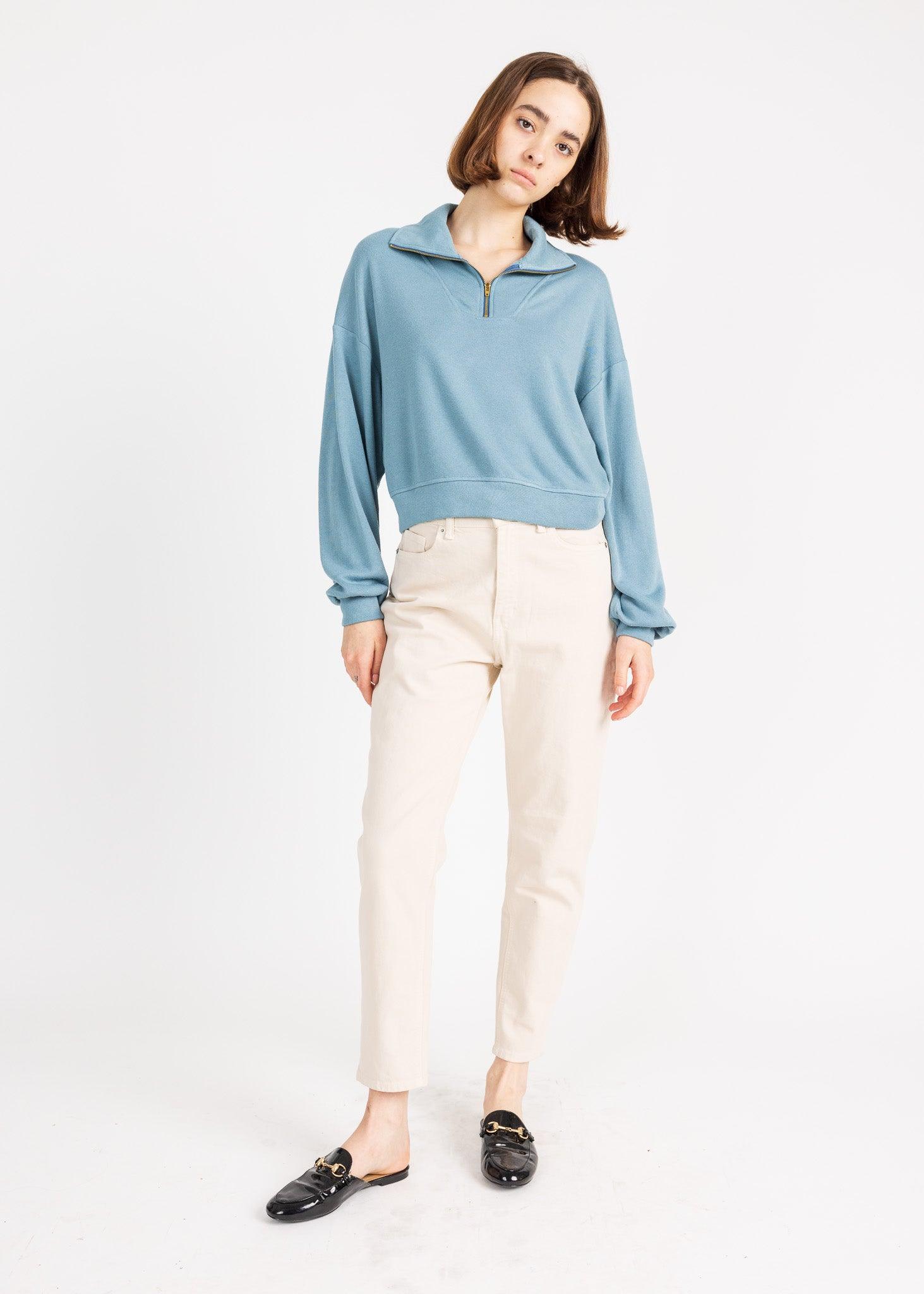 A woman wearing a blue zip-up sweater and cream-colored pants poses against a white background.