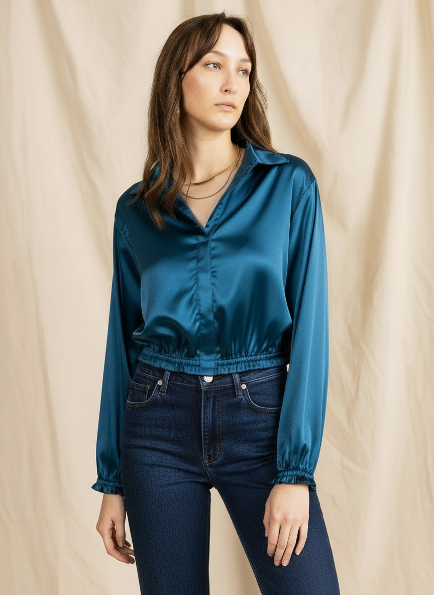 A person wearing a beige satin top with long sleeves, a collared neck, and a cropped fit with ruffled cuffs. The person is also wearing blue jeans.