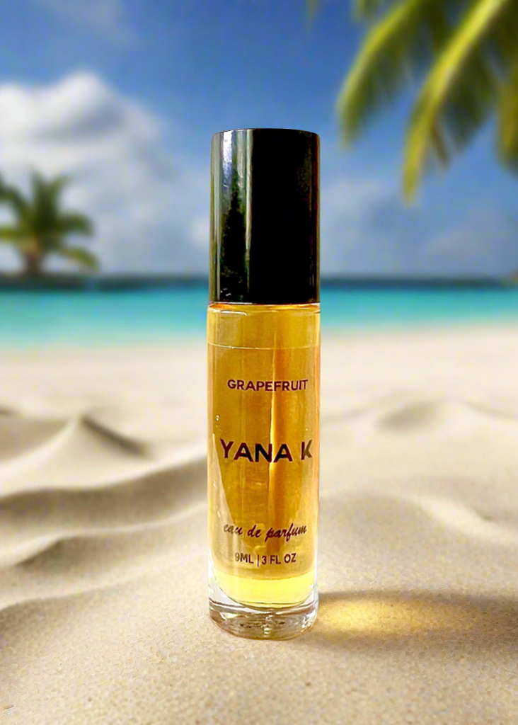 YANA K grapefruit perfume bottle on a sandy beach with palm trees and blue sky.