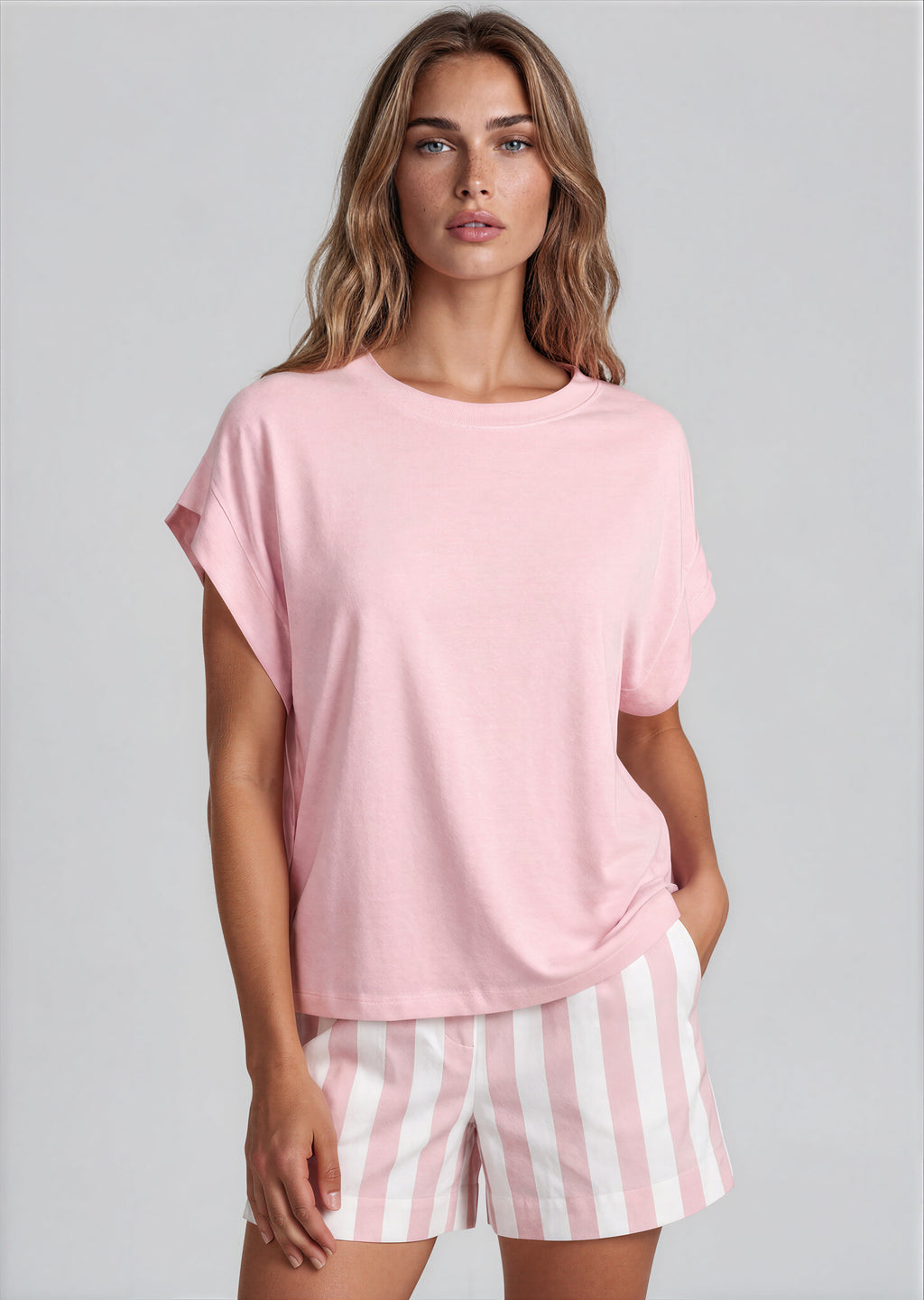 A woman modeling a light pink short-sleeve crewneck t-shirt and pink and white vertically striped shorts against a solid light gray background.
