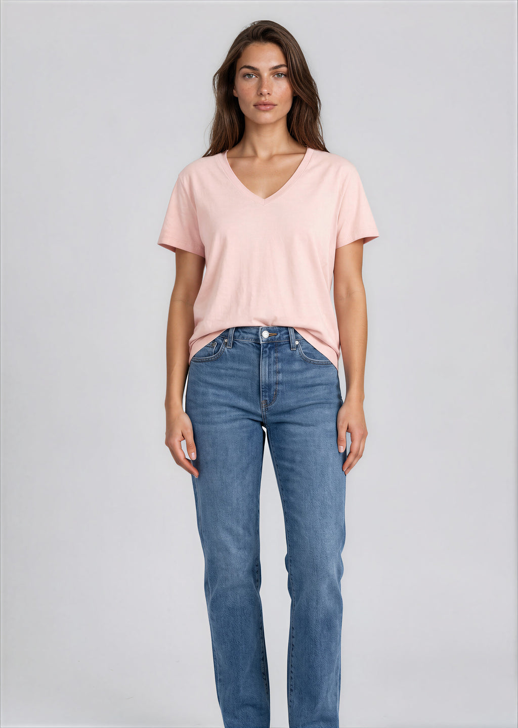 A woman modeling a light pink v-neck t-shirt and blue jeans against a plain white studio background.