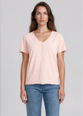 A woman modeling a light pink short-sleeve V-neck t-shirt tucked into blue jeans, photographed against a solid light gray studio background.