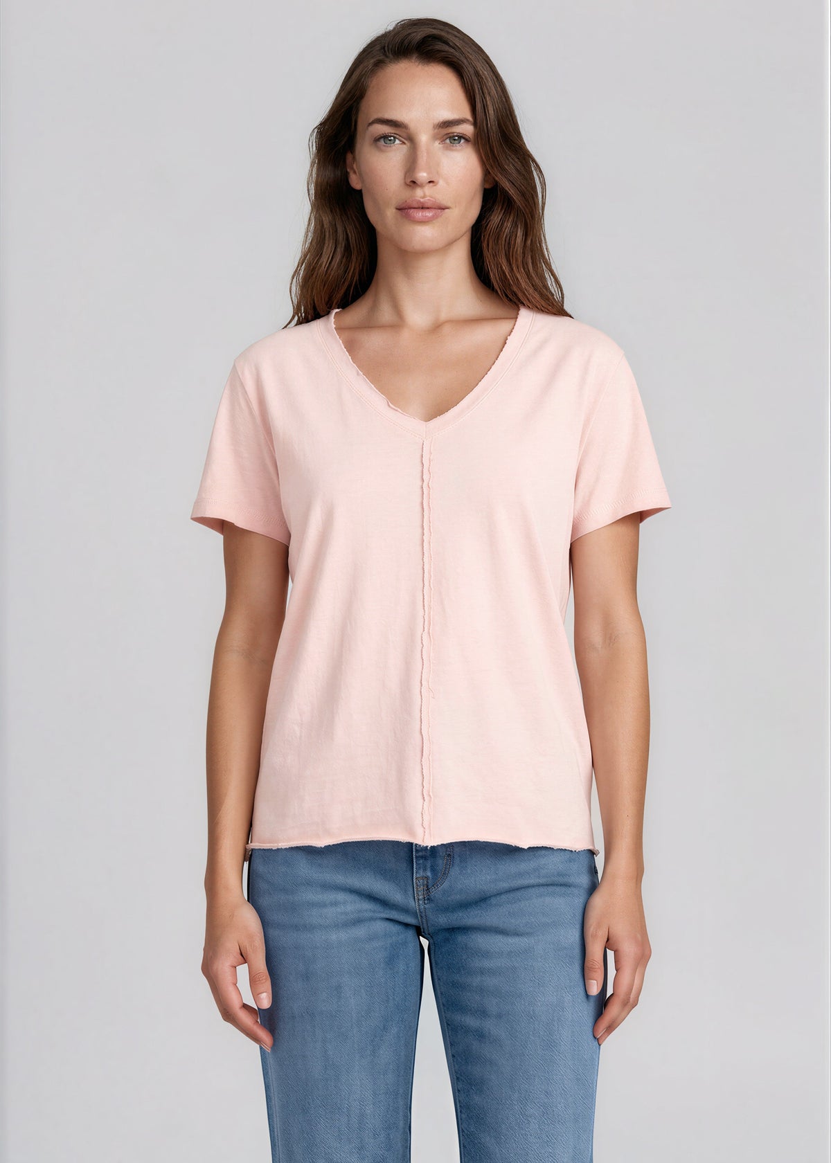 A woman modeling a light pink short-sleeve V-neck t-shirt tucked into blue jeans, photographed against a solid light gray studio background.