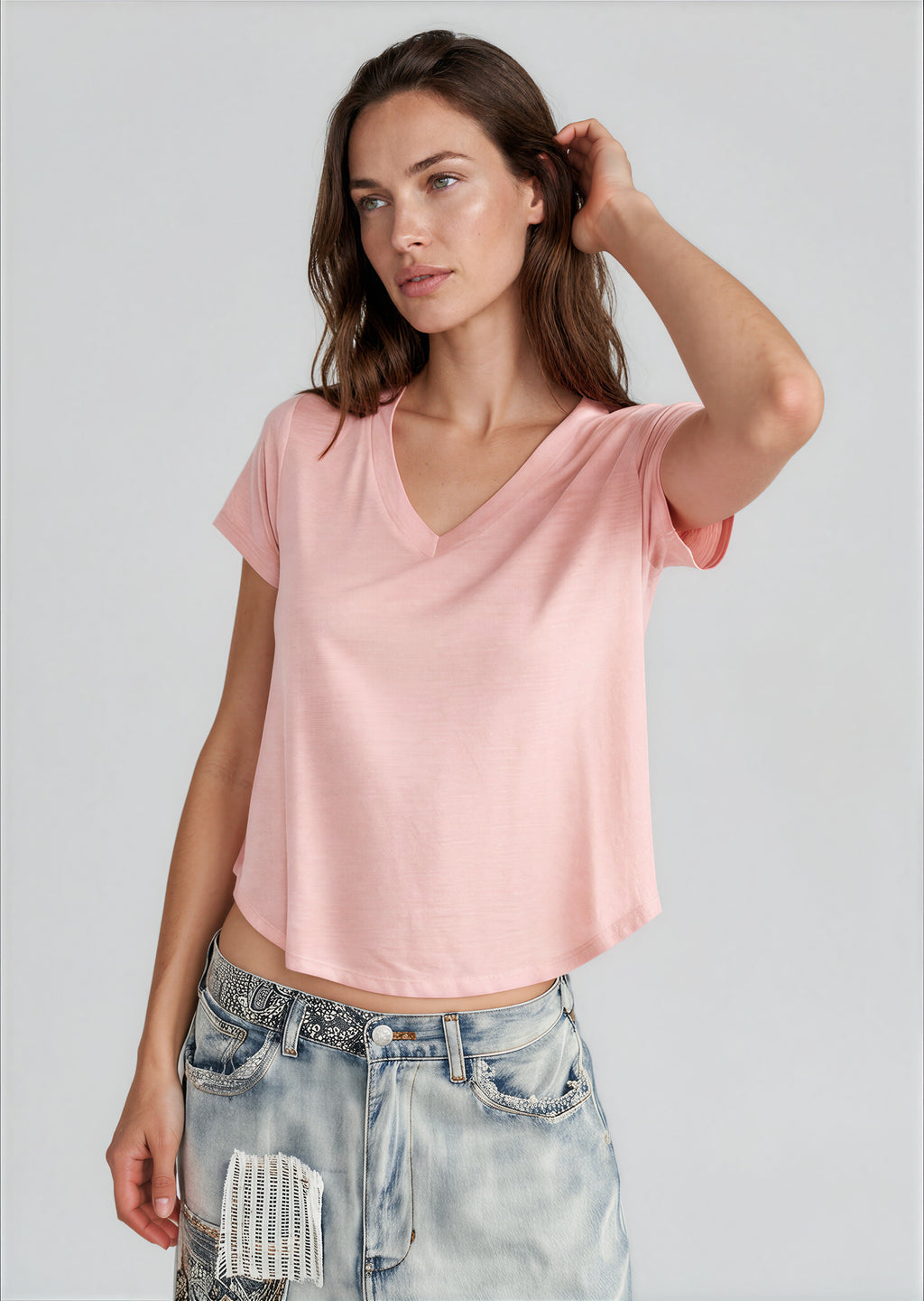 A woman wearing a light pink short-sleeve V-neck crop top and light-wash distressed denim jeans, standing against a solid light gray studio background.
