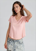 A woman wearing a light pink short-sleeve V-neck crop top and light-wash distressed denim jeans, standing against a solid light gray studio background.